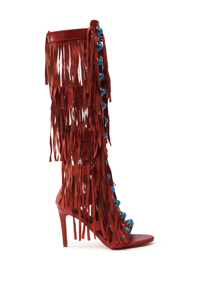 The AZALEA WANG AMADEO RED SANDAL is a gladiator-style, high-heeled sandal with cascading red fringe layers on the sides and blue spherical accents on the front. It features an open toe design and a stiletto heel.