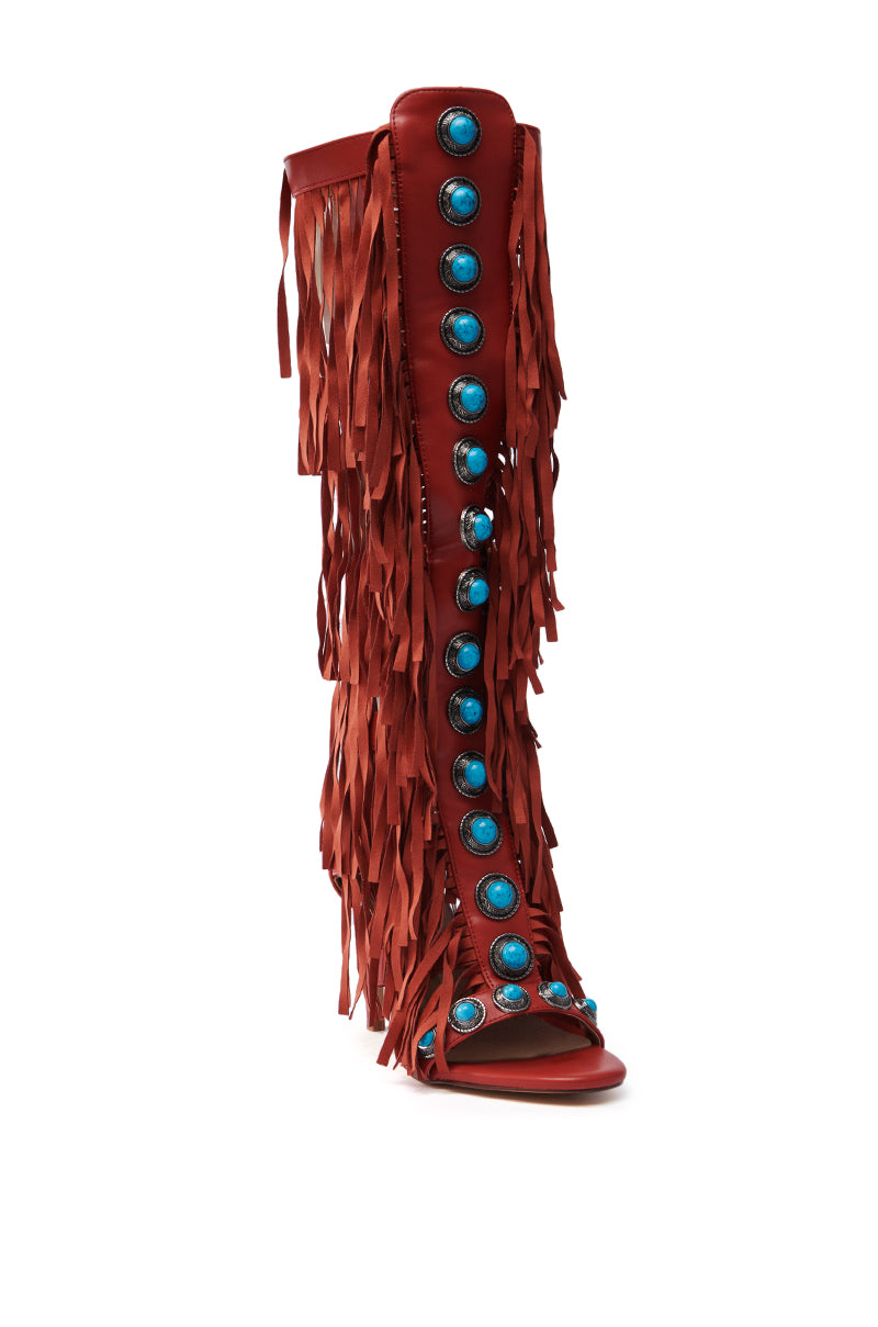 The AZALEA WANG AMADEO RED SANDAL is a tall, gladiator-style open-toe boot made of vegan leather, featuring turquoise gemstone embellishments on the front and dramatic red fringe detailing on the sides for a bold and unique look.