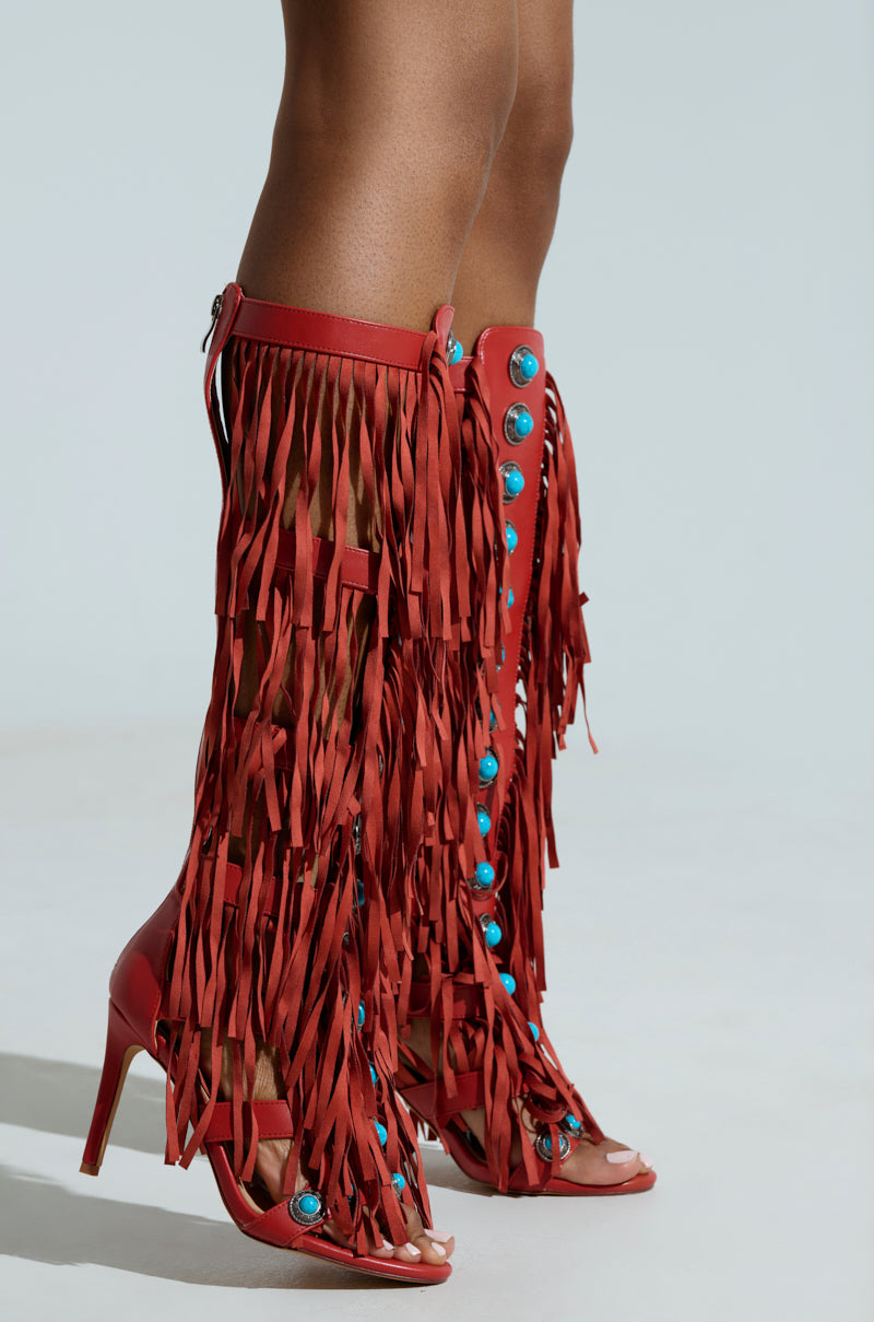 Wearing the AZALEA WANG AMADEO RED SANDAL, a person models knee-high, red high-heeled boots with a gladiator silhouette. Turquoise gemstones and long fringe accent the peep-toe design, and a back zipper closure adds chic flair.