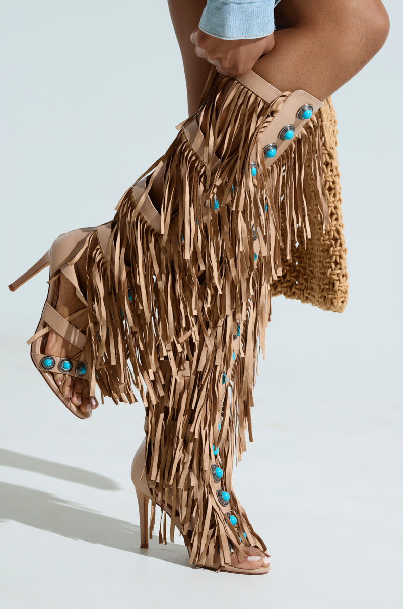A person dons the AZALEA WANG AMADEO TAN SANDAL, a high-heeled, knee-high beige boot with stiletto heels, long fringes, turquoise accents, and open toes. Paired with a woven bag against a neutral backdrop.
