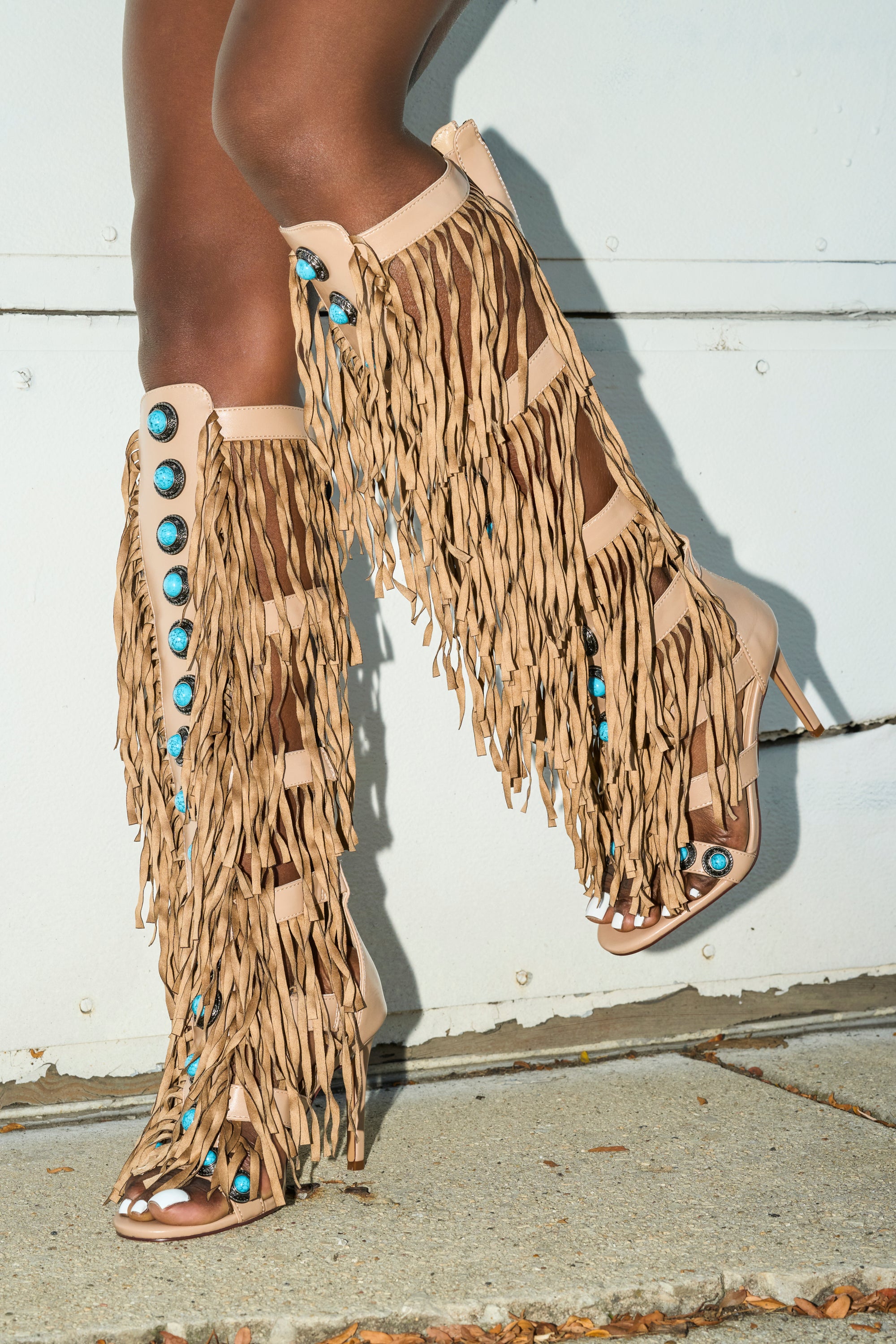 A person stands on a pavement against a white wall wearing the AZALEA WANG AMADEO TAN SANDAL—knee-high gladiator sandals with stiletto heels, tan fringe, and turquoise studs.