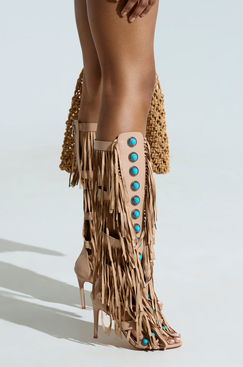 A person dons the AZALEA WANG AMADEO TAN SANDAL, knee-high gladiators with fringe and turquoise accents, while standing on a light surface and holding a textured woven handbag.