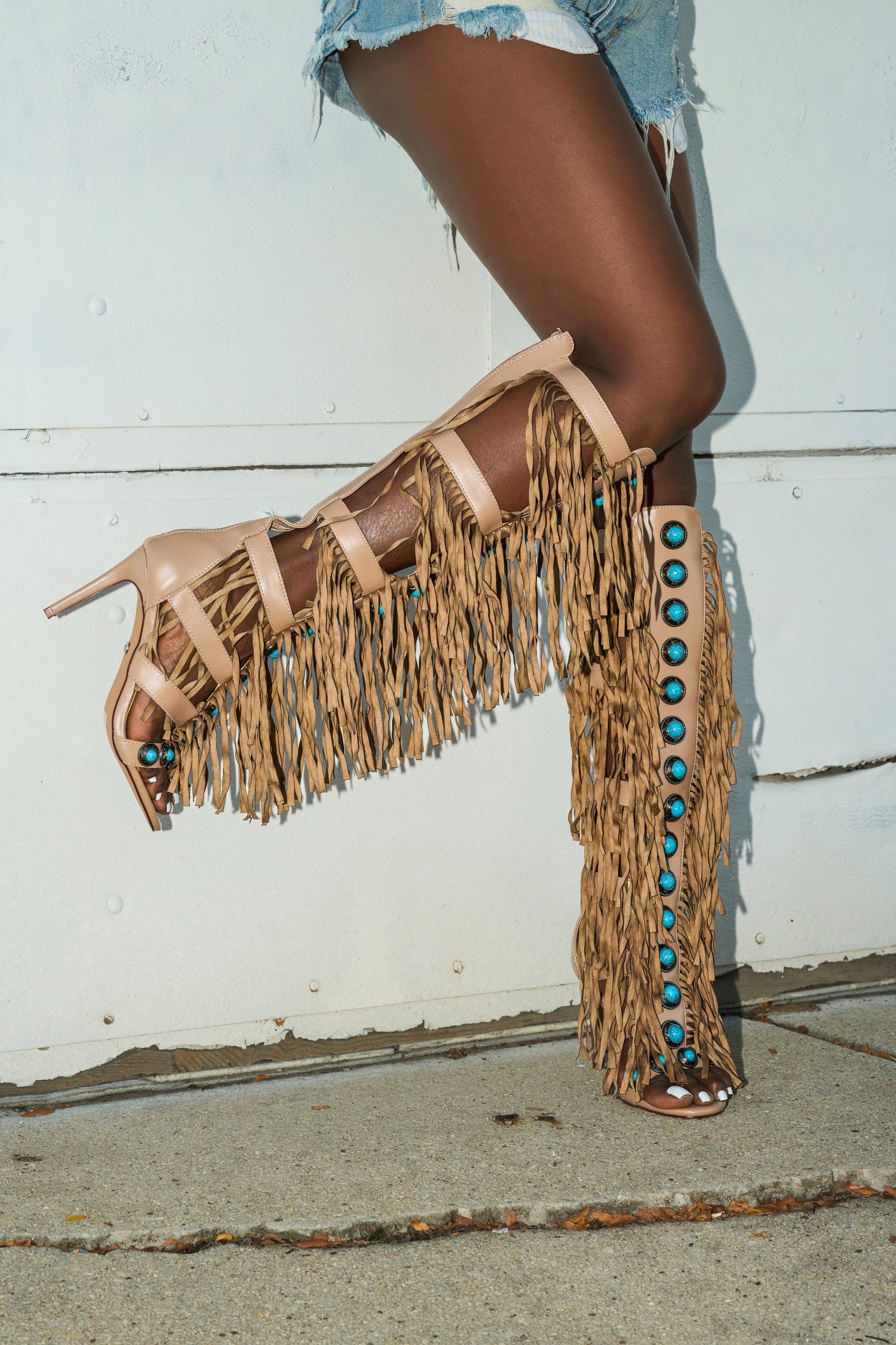Wearing denim shorts and the AZALEA WANG AMADEO TAN SANDAL—tan, high-heeled, knee-high sandals with fringe and turquoise bead accents—this person stands on a concrete surface.