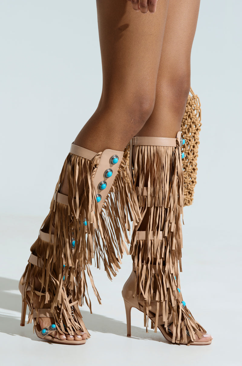 A close-up features AZALEA WANG AMADEO TAN SANDAL, a gladiator design with long tan faux leather fringe and turquoise button accents, extending from the top to mid-calf. The person is also holding a woven textured handbag.