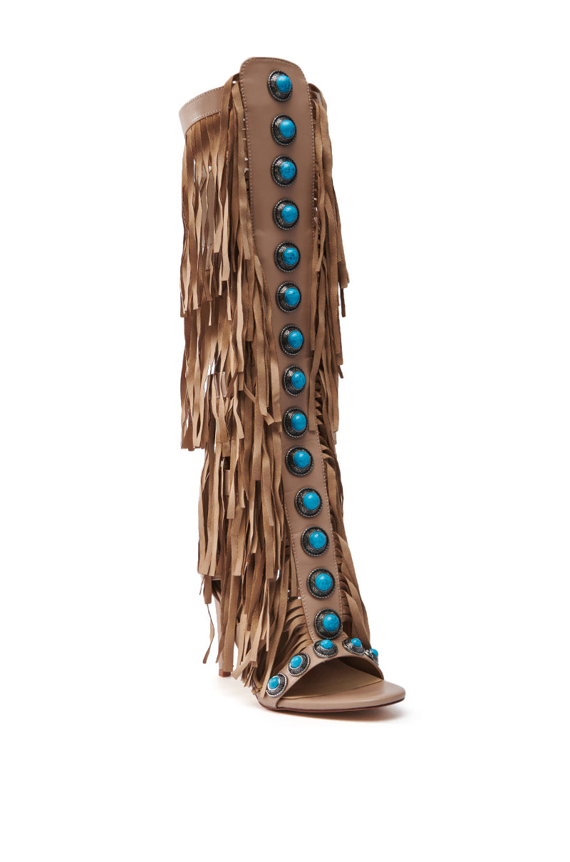 The AZALEA WANG AMADEO TAN SANDAL is a knee-high open-toe boot in tan faux leather, featuring fringe on the sides and front. A striking row of turquoise stones adorns it from top to bottom, making these stiletto heel boots a bold statement piece.