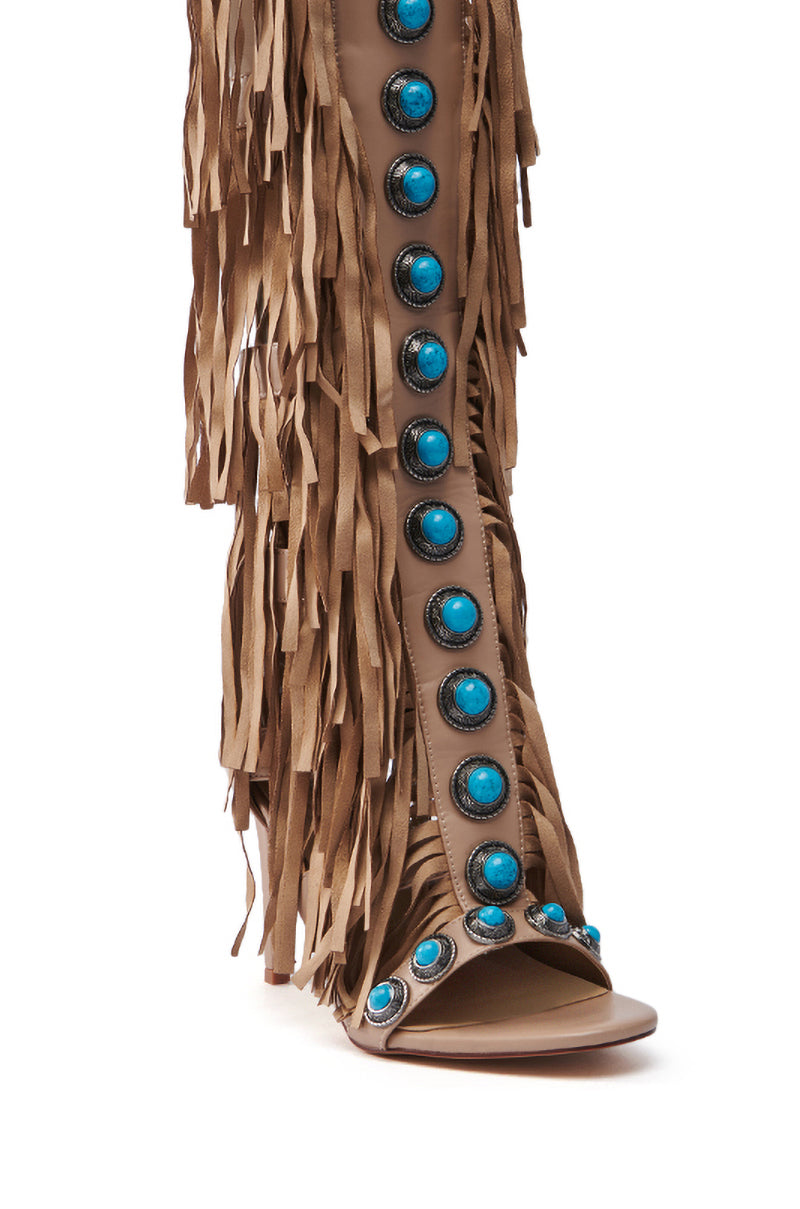 The AZALEA WANG AMADEO TAN SANDAL is a knee-high gladiator sandal with brown fringe along the sides and turquoise stones on a beige faux leather strip, featuring an open-toe design for a chic boho vibe.