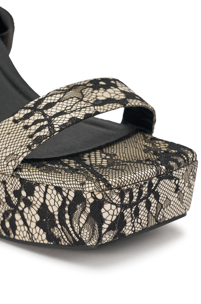 Close-up of AKIRA GOLD AMBROISINE black lace strappy chunky sandals with a high block heel and wide toe strap, set against a beige background. 8