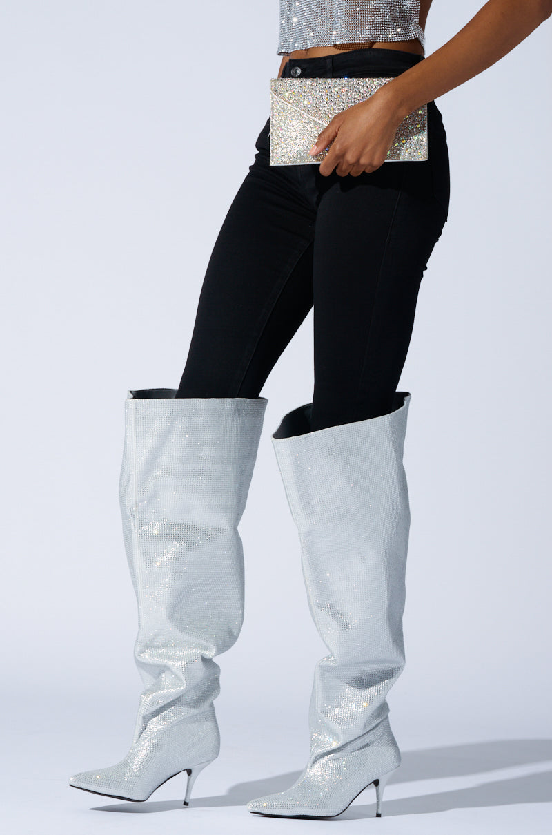 A person wearing the AZALEA WANG AMBY SILVER BUCKET BOOT, featuring sparkling rhinestone details, knee-high design, pointed toe, and a small heel is paired with black pants and holding a glittery clutch. The ensemble is styled to achieve a fashionable or glamorous look.