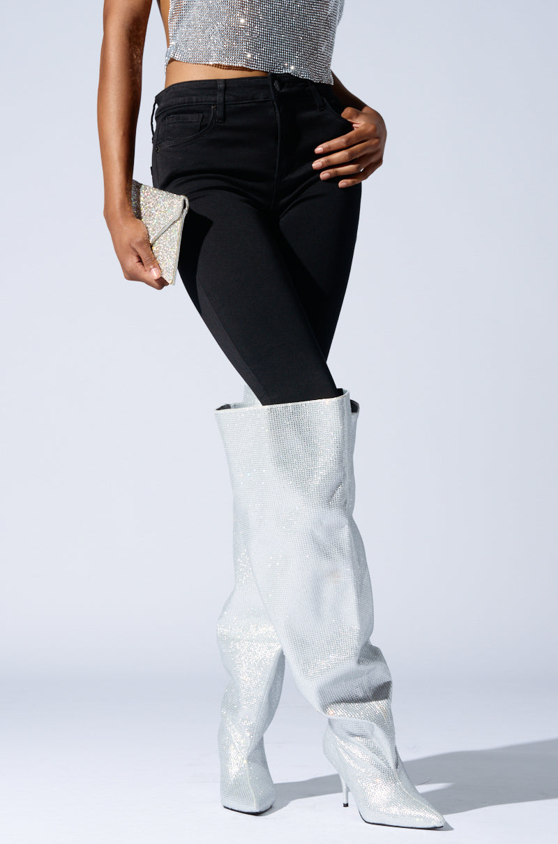 A person wearing black pants and a glittery silver top is showcasing the AZALEA WANG AMBY SILVER BUCKET BOOT, featuring knee-high design with rhinestone detailing, while holding a sparkly silver clutch against a plain light gray background.