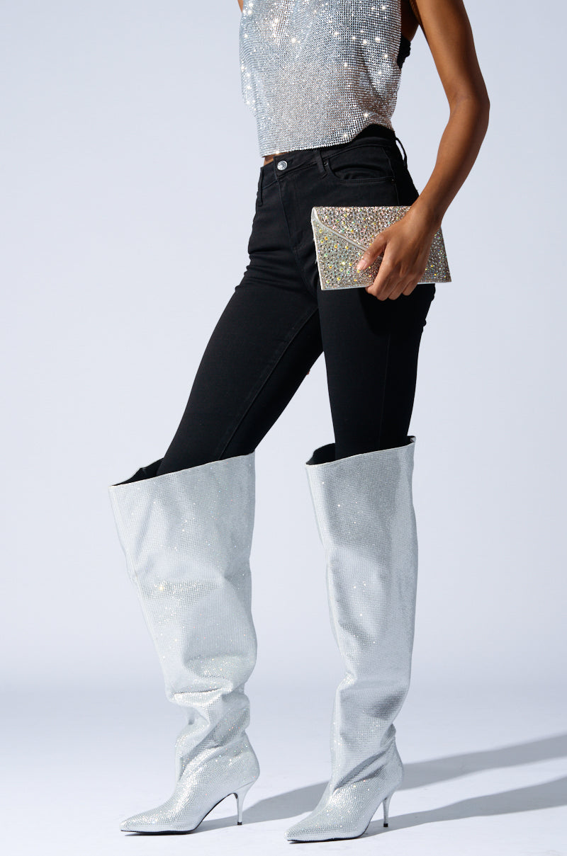 A person wearing a shiny silver top and black jeans confidently dons the AZALEA WANG AMBY SILVER BUCKET BOOT, featuring tall silver glittery thigh high design with intricate rhinestone detailing, while holding a sparkling clutch. The plain background highlights the outfit's reflective textures.