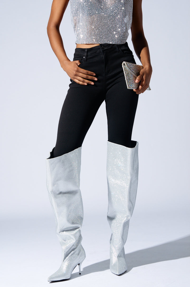 A person wearing a sparkly silver sleeveless top, black fitted pants, and the AZALEA WANG AMBY SILVER BUCKET BOOTS with rhinestone detailing. They are holding a small, shiny clutch bag against a plain, light gray background.