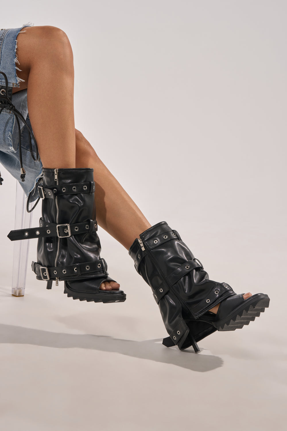 A person in distressed denim shorts sits cross-legged, showcasing the AZALEA WANG AMES Black PU Hardware Embellished Stiletto Bootie with a sleek faux leather finish and bold hardware accents. 7