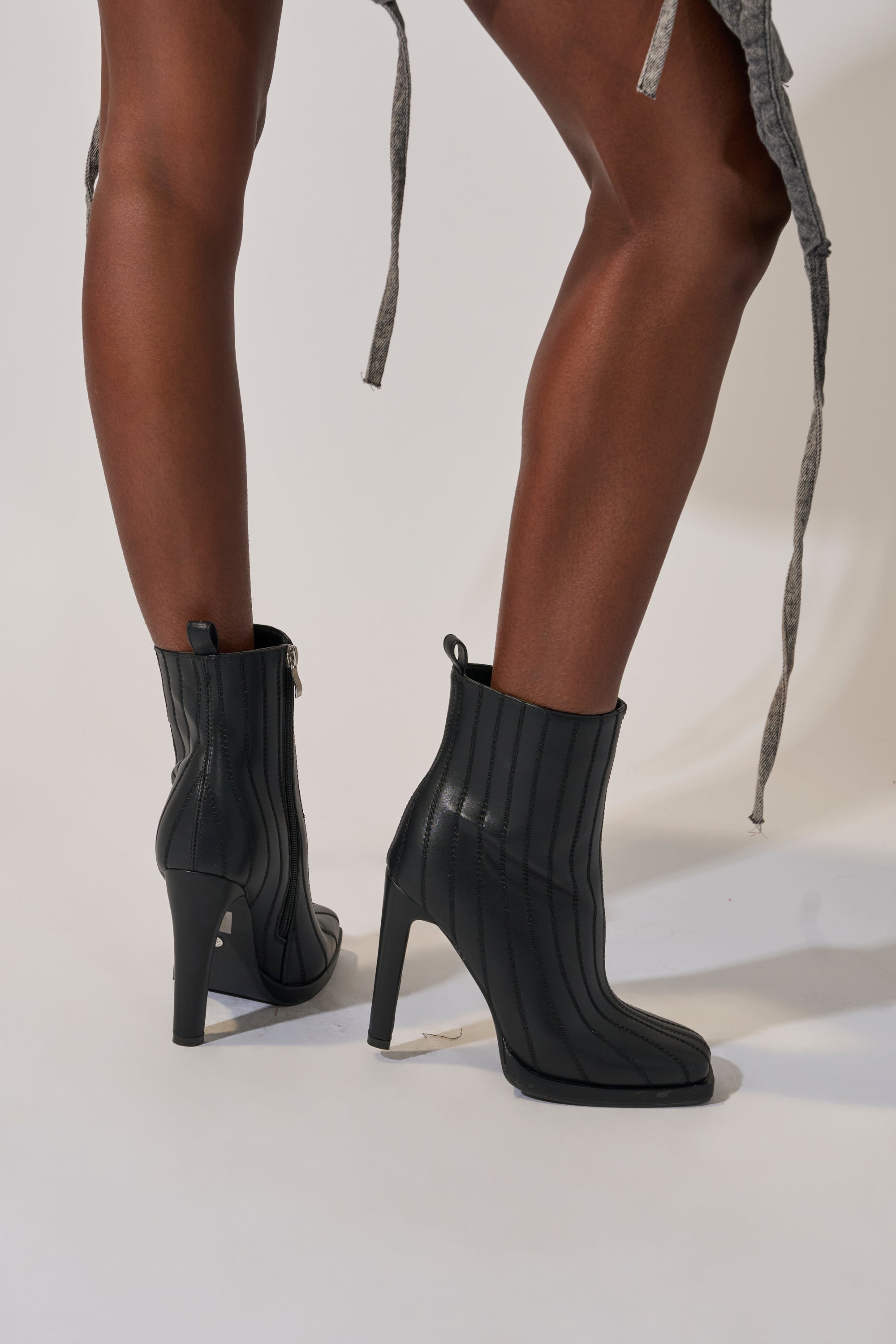 Someone stands on a light-colored surface wearing the AZALEA WANG AMHERST BLACK PU BOOTIE with vertical stitching and a chunky heel, paired with a gray textured skirt that has loose fabric ties.