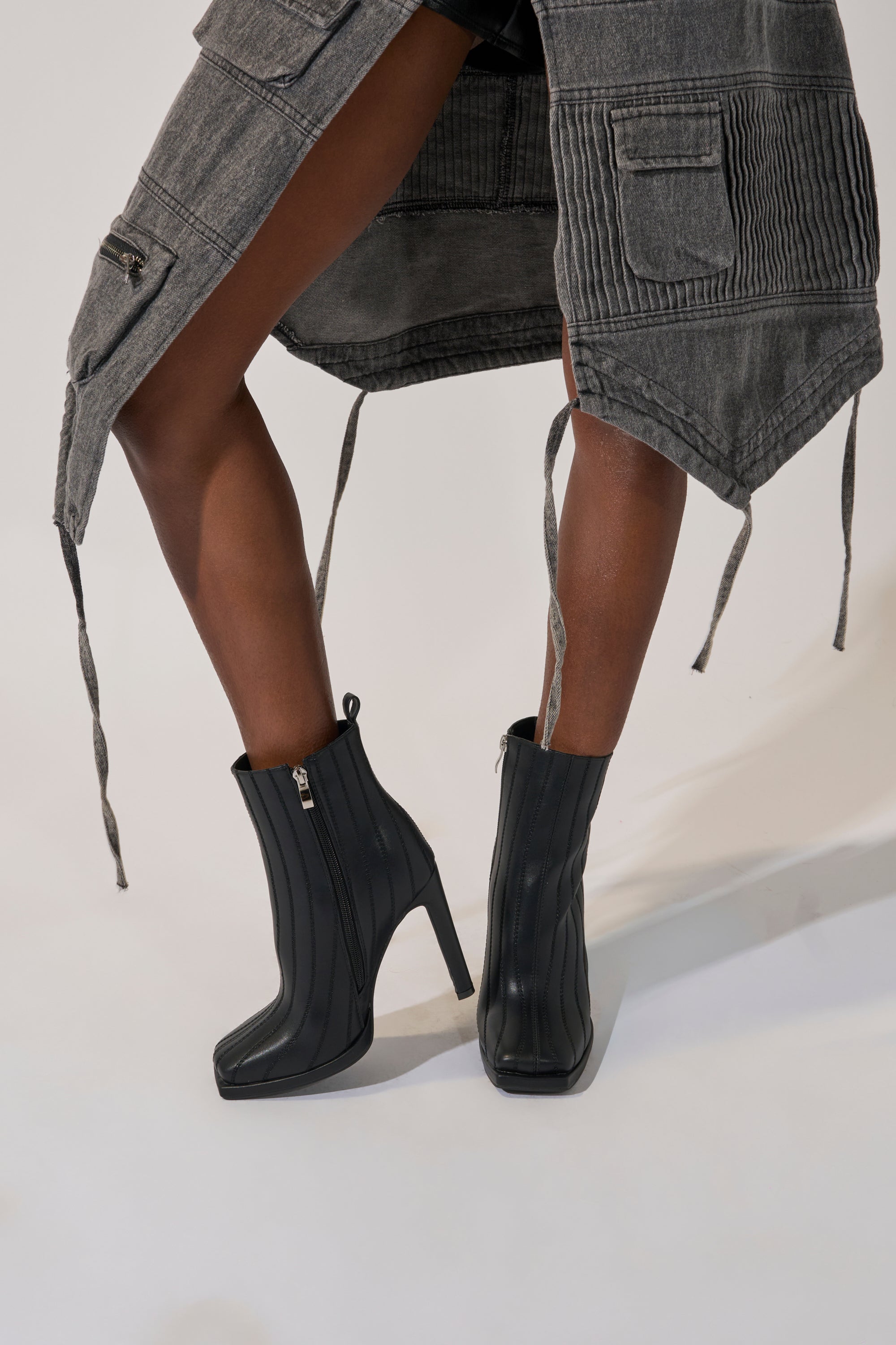 A person wearing a long, gray, pocketed coat and the AZALEA WANG AMHERST BLACK PU BOOTIE stands with one leg crossed in front of the other against a plain background.