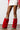 Wearing a white skirt and a red textured jacket, the person completes their ensemble with the AZALEA WANG AMOURA RED FUR AND SNAKE BOOT—knee-high, red furry boots showcasing faux snakeskin accents at the top and toes—as they stand indoors on a wooden floor.