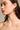 A person with long brown hair is shown from the shoulders up, looking to the side. They're wearing DRIPPY EARRINGS, faux gold with an abstract design. The background is plain and neutral.