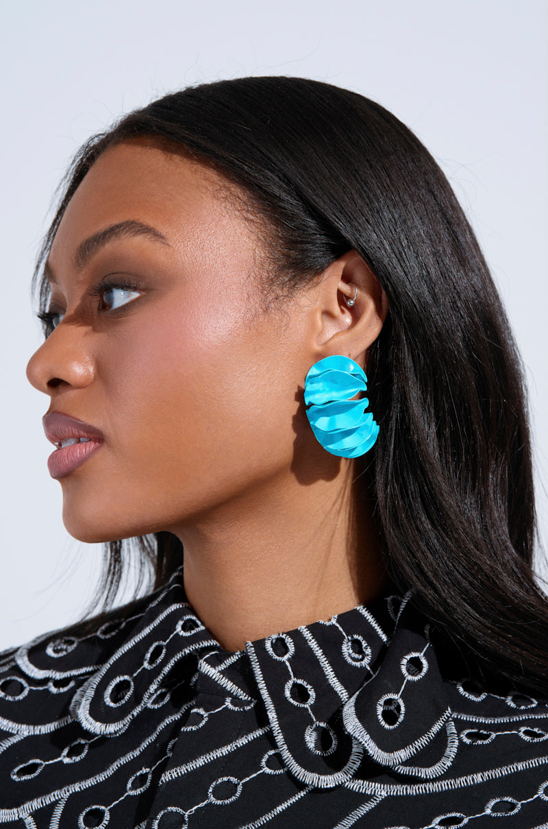 A woman with long black hair is wearing a black and white patterned top, paired with FANNED OUT EARRING in bright sky blue metal. She looks to the side against a plain background.