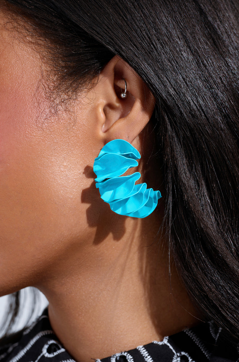 A person wears a large, sky blue "FANNED OUT EARRING," its metal spiral shape creating a vibrant contrast with their dark hair and skin. A small hoop adorns their upper ear, perfectly complementing the striking black and white striped outfit.