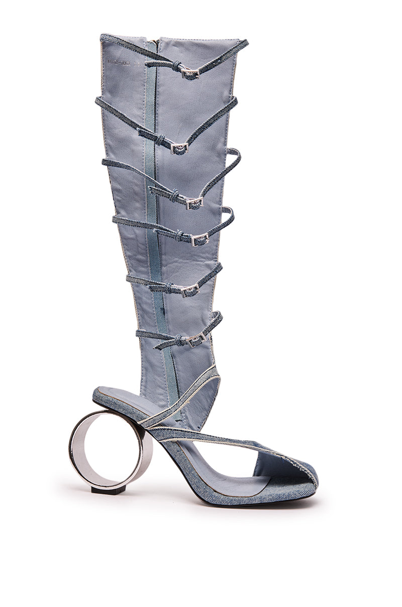 The AZALEA WANG ANDREE DENIM GLADIATOR SANDAL features tall, light blue denim straps and a distinctive sculptural circular metal heel, shown against a white background.