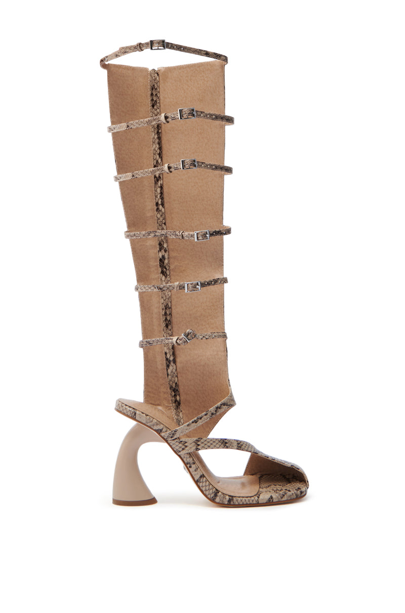 The AZALEA WANG ANDRIUS SNAKE CHUNKY GLADIATOR SANDAL is a stylish, knee-high gladiator sandal featuring a unique curved heel and multiple straps in a striking snakeskin pattern. Its faux leather design adds elegance, and the open-toe style provides a trendy touch.