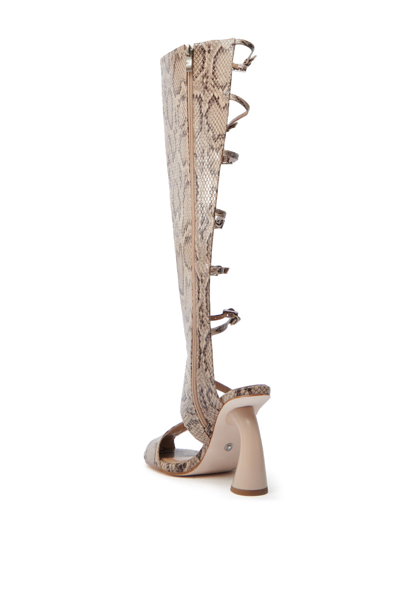 The AZALEA WANG ANDRIUS SNAKE CHUNKY GLADIATOR SANDAL features a side view of a high heel with an open strappy shaft, striking snakeskin pattern, unique curved beige heel, and faux leather. It has multiple straps and a side zipper for easy fastening.