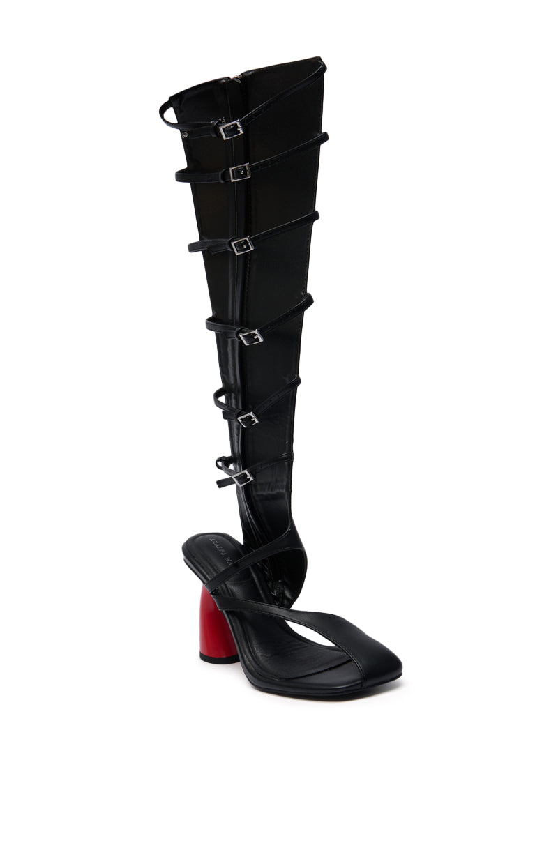 The AZALEA WANG ANDRIUS BLACK WITH RED HEEL GLADIATOR SANDAL features a black faux leather design, a striking red chunky heel, a tall structured gaiter, and multiple silver buckles up the shaft.