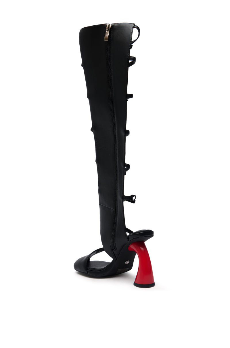 The AZALEA WANG ANDRIUS BLACK WITH RED HEEL GLADIATOR SANDAL is a black, open-toe, knee-high faux leather boot featuring cutouts, buckle accents, bold exposed sides, a chunky red heel, and a side zipper for a modern gladiator look.