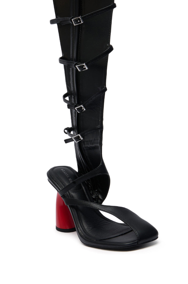 The AZALEA WANG ANDRIUS BLACK WITH RED HEEL GLADIATOR SANDAL features black faux leather, a bold red chunky heel, and multiple thin buckled straps running up the leg, shown styled over a black stocking.