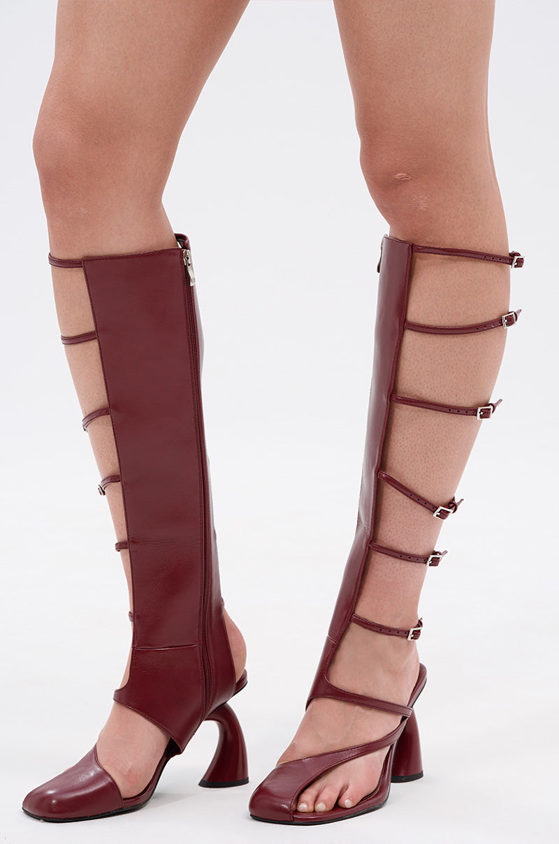 A model wears the AZALEA WANG ANDRIUS BURGUNDY GLADIATOR SANDAL—knee-high, open-toe faux leather boots with side straps, buckles, and a curved chunky heel—standing against a plain white background.