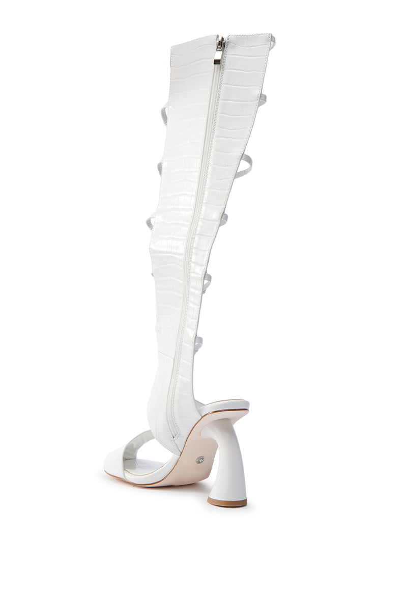 The AZALEA WANG ANDRIUS WHITE CROC CHUNKY GLADIATOR SANDAL is a striking knee-high, white high-heeled boot made from glossy croc-embossed faux leather. It features an open-toe, strappy design, a unique sculpted heel, and a long back zipper.