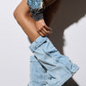 A person dressed in oversized denim leg warmers and shorts featuring a detailed blue paisley design leans against a wall. They are wearing the AZALEA WANG ANGELA FOLD OVER PLATFORM BOOT IN DENIM, which are scrunched for a trendy, laid-back style. The scene is set on a light wooden floor.