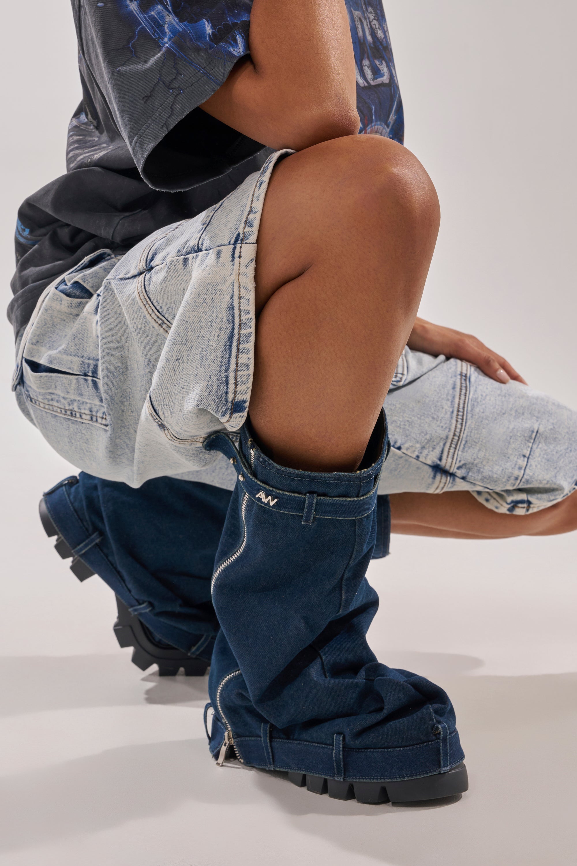 A person squats, showcasing the AKIRA GOLD ANGELA FOLD OVER PLATFORM BOOT IN DARK DENIM with oversized light-wash denim shorts over dark jeans. Only their legs and part of their torso appear against a plain background. 1