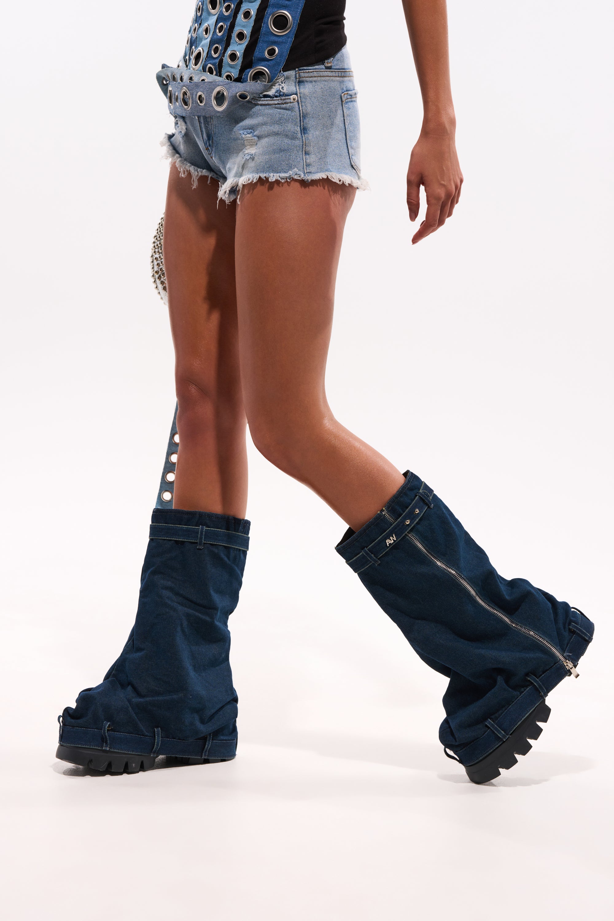 Wearing distressed denim shorts, a studded belt, and AKIRA GOLD ANGELA FOLD OVER PLATFORM BOOT IN DARK DENIM with a chunky lug sole, a person walks against a plain white background. Only the lower body is visible. 0