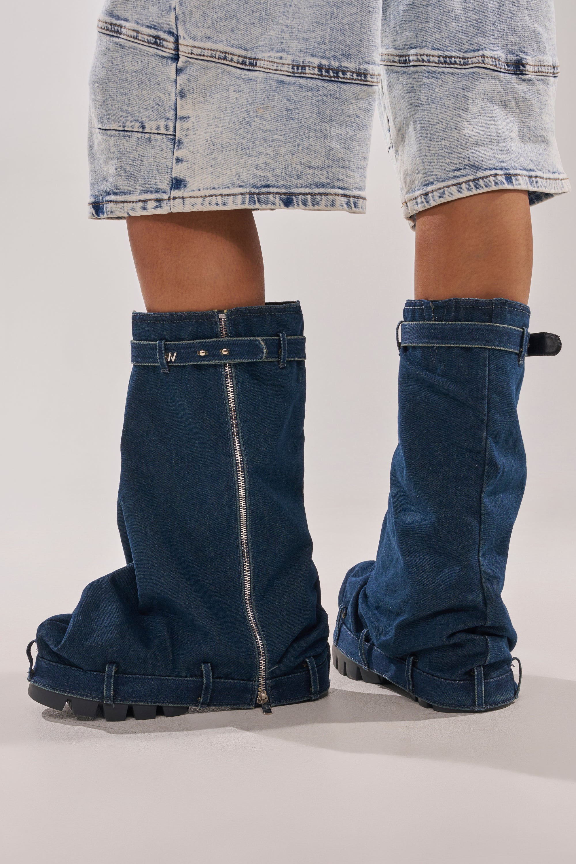 A person stands on a light background wearing light-washed, knee-length denim shorts and AKIRA GOLD ANGELA FOLD OVER PLATFORM BOOTS IN DARK DENIM, featuring a chunky lug sole, zippers, and straps. 2