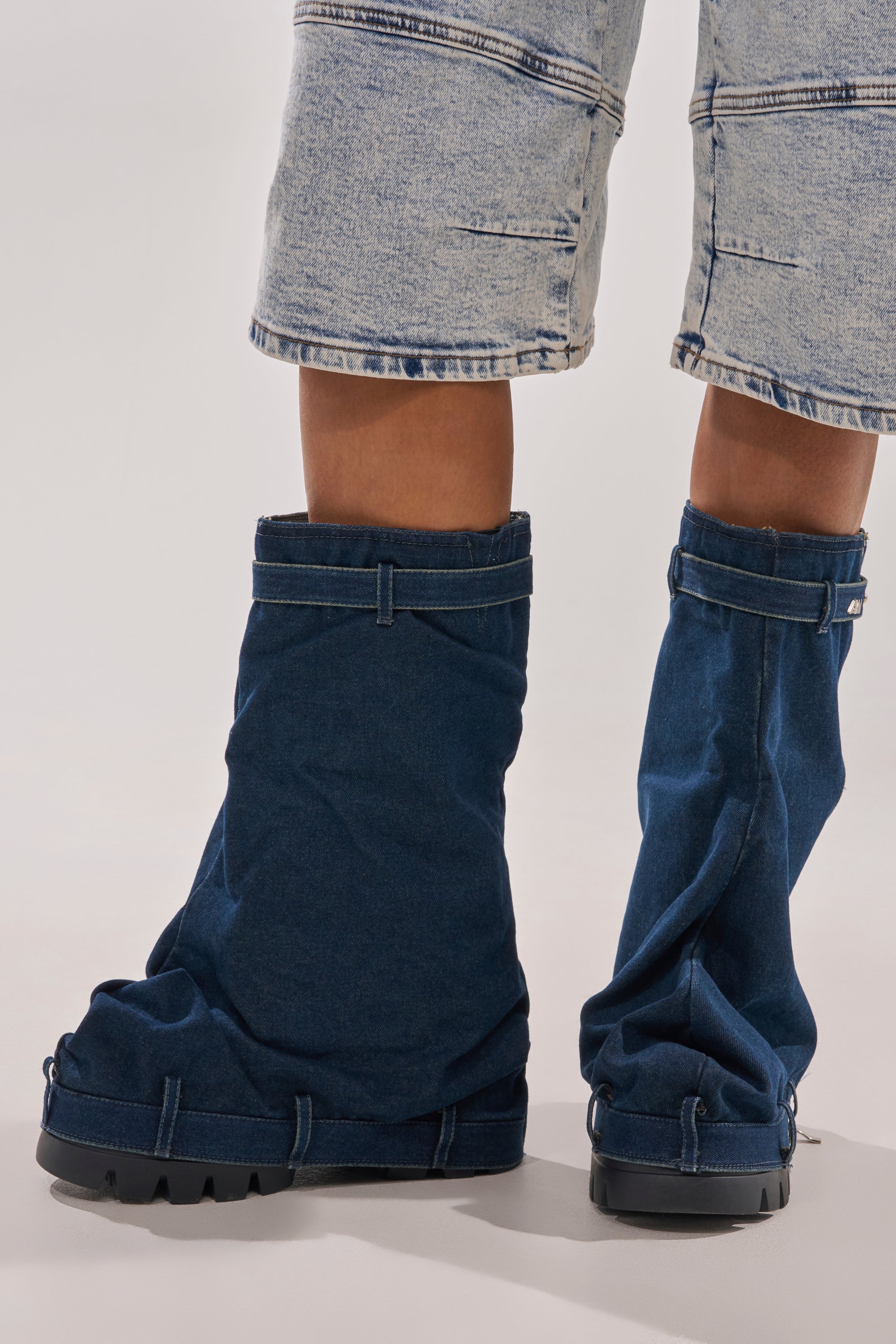 A model stands on a plain white background wearing light-wash cutoff jeans and the AKIRA GOLD ANGELA FOLD OVER PLATFORM BOOT IN DARK DENIM, featuring an oversized slouchy silhouette and chunky black soles. 8