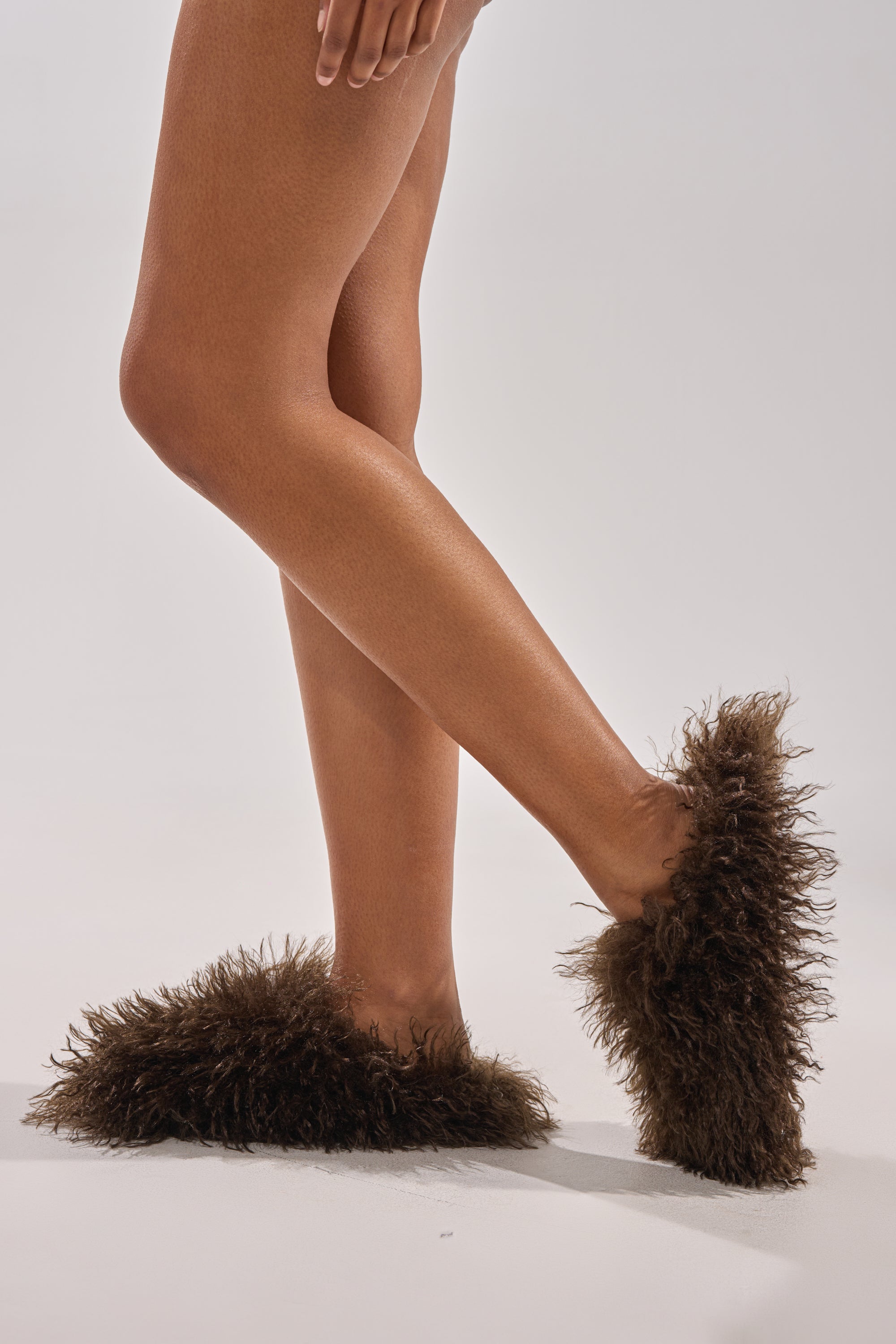 A person with crossed legs wears AKIRA ANGOL CHOCOLATE SHAGGY FLAT SLIPPERS, standing on a light neutral background. 1