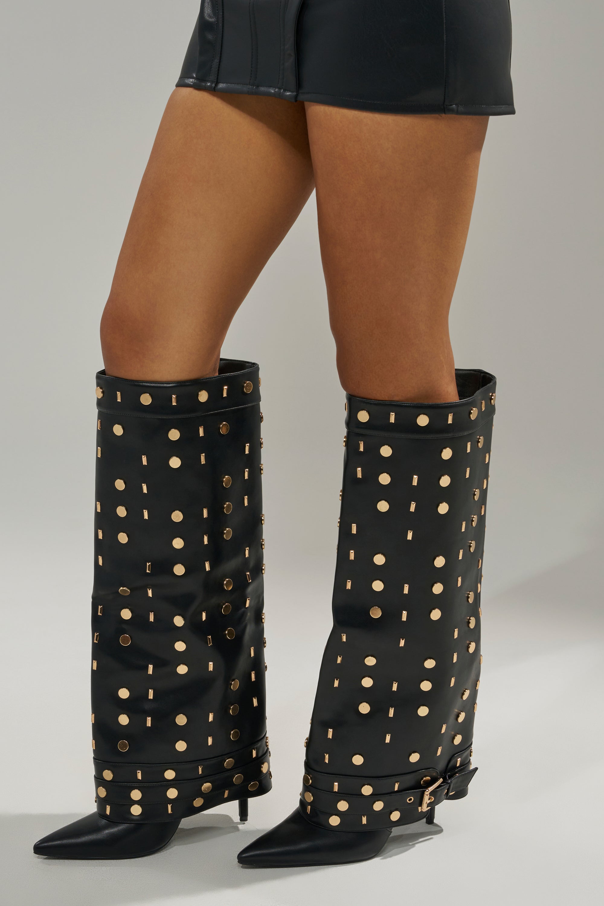 A person stands on a plain light background wearing the AZALEA WANG ANISSA Black Multi Gold Hardware Stiletto Boot with black faux leather shorts.