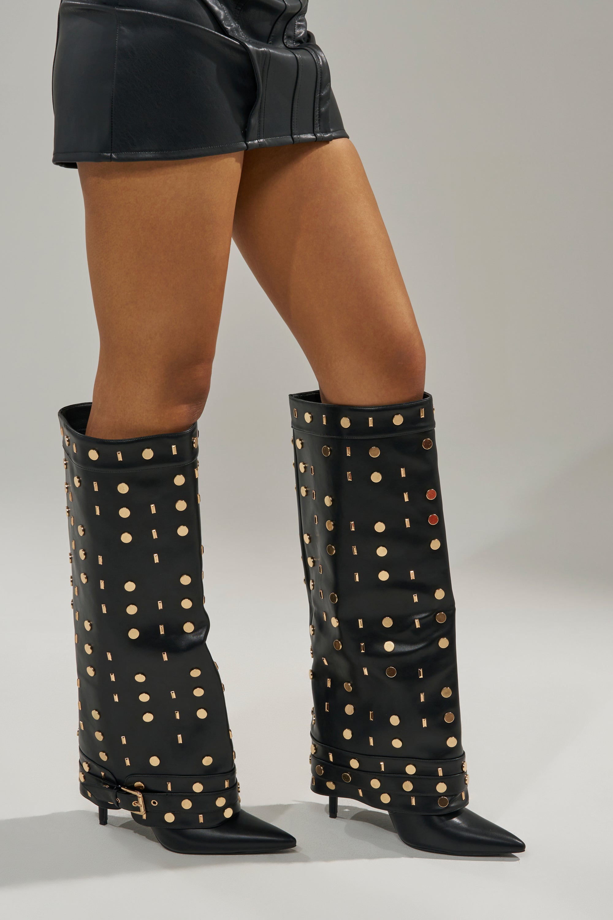Wearing AZALEA WANG ANISSA Black Multi Gold Hardware Stiletto Boots with black leather shorts, a person stands against a plain gray background.
