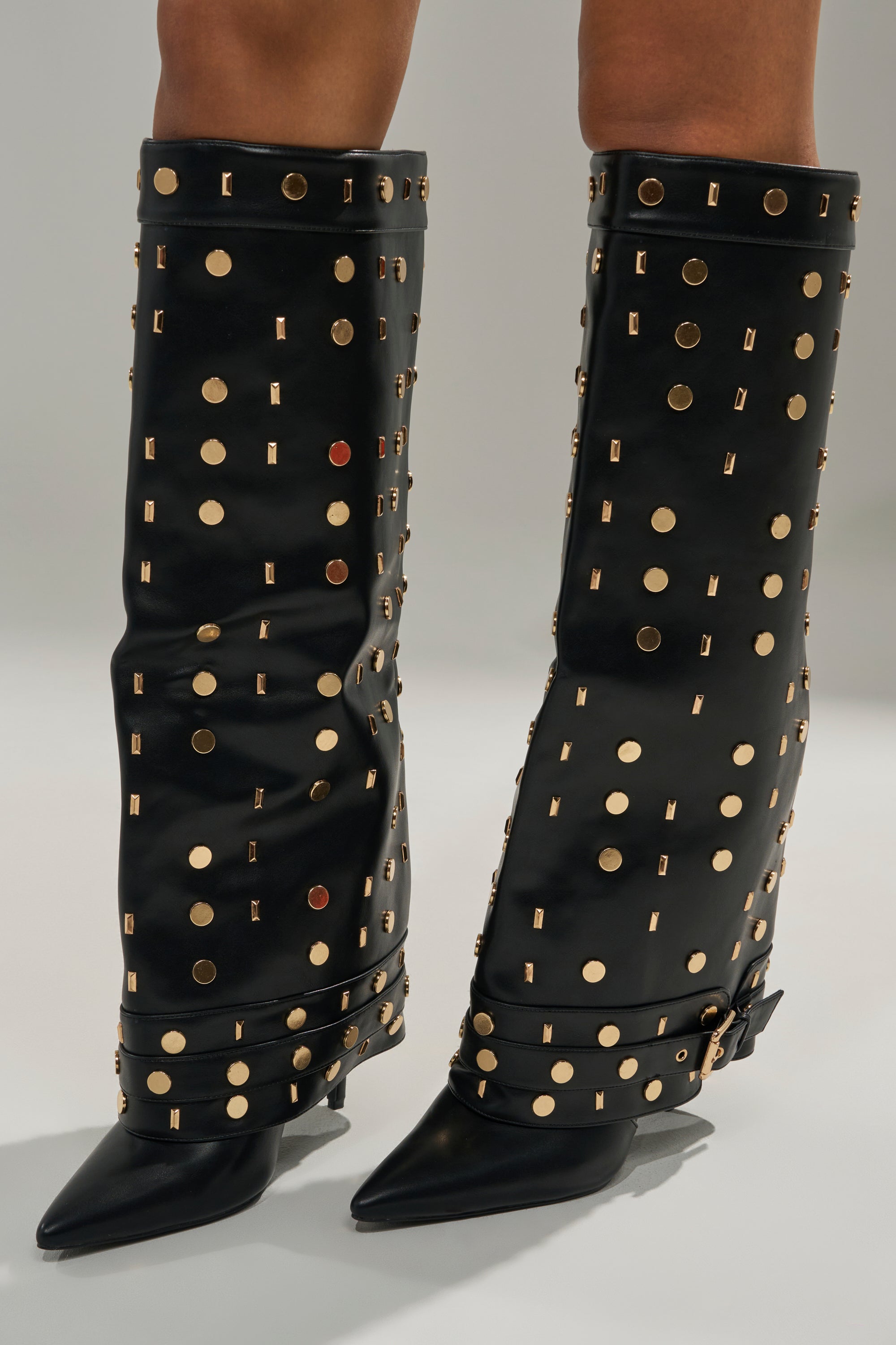 Close-up of AZALEA WANG ANISSA Black Multi Gold Hardware Stiletto Boots—black faux leather, knee-high, pointed toes, gold studs and circular metal accents, buckled ankle straps. Worn with bare legs.