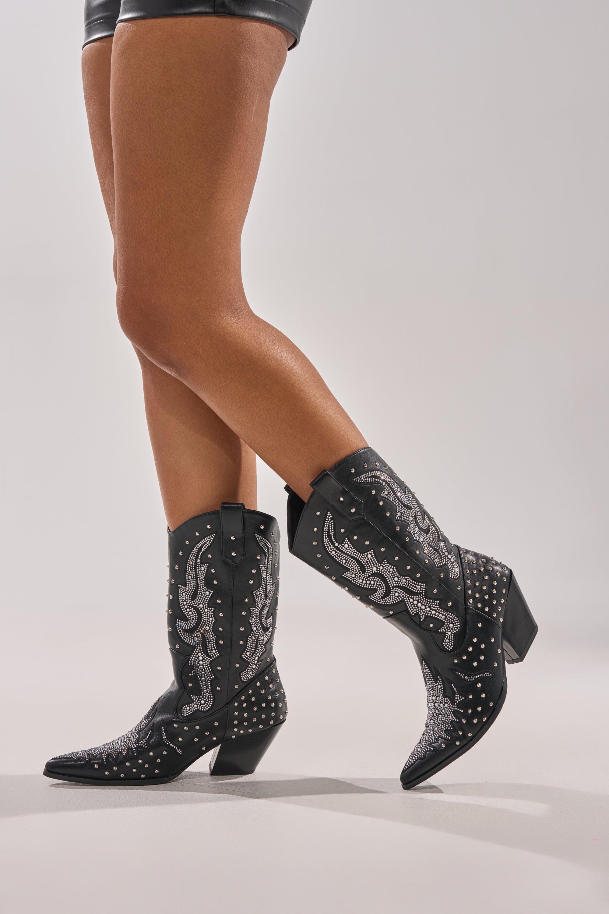 A person stands on a light grey background, wearing only black leather shorts and the AZALEA WANG ANKUS BLACK WESTERN RHINESTONE BOOT with mid heels. Only their legs and feet are visible.
