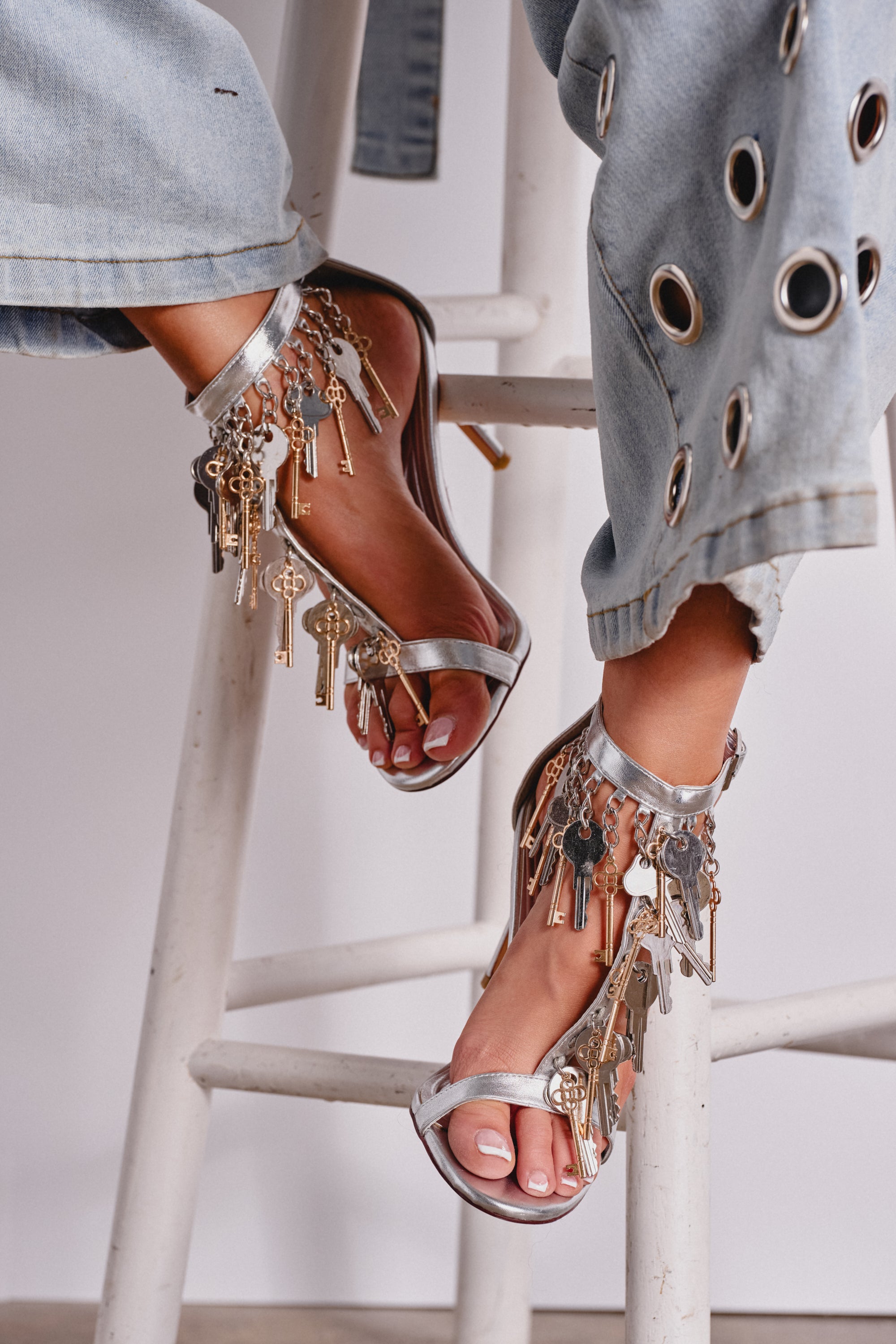Close-up of a person wearing AKIRA GOLD ANNIKI SILVER KEY EMBELLISHED NOVELTY STILETTO SANDALS with light blue jeans featuring large metal grommets, seated on a white wooden stool. 0