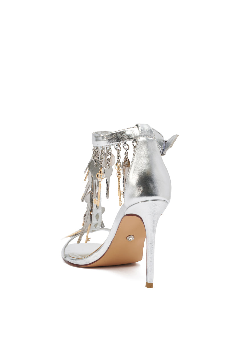 The AKIRA GOLD ANNIKI Silver Key Embellished Novelty Stiletto Sandal features metallic faux leather, an ankle strap, and a heel adorned with dangling key and lock charms, shown from the back on a white background. 6