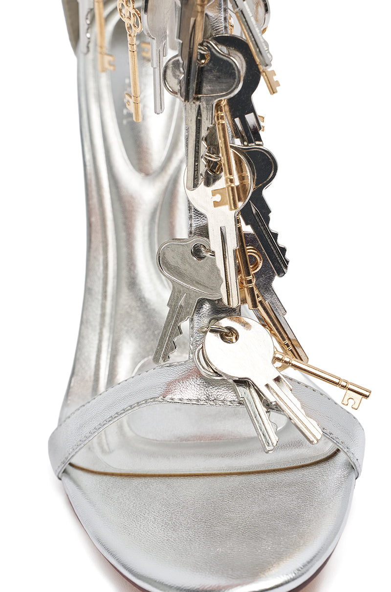 A close-up of the AKIRA GOLD ANNIKI Silver Key Embellished Novelty Stiletto Sandal features a metallic faux leather design with multiple decorative keys hanging from the straps. 7