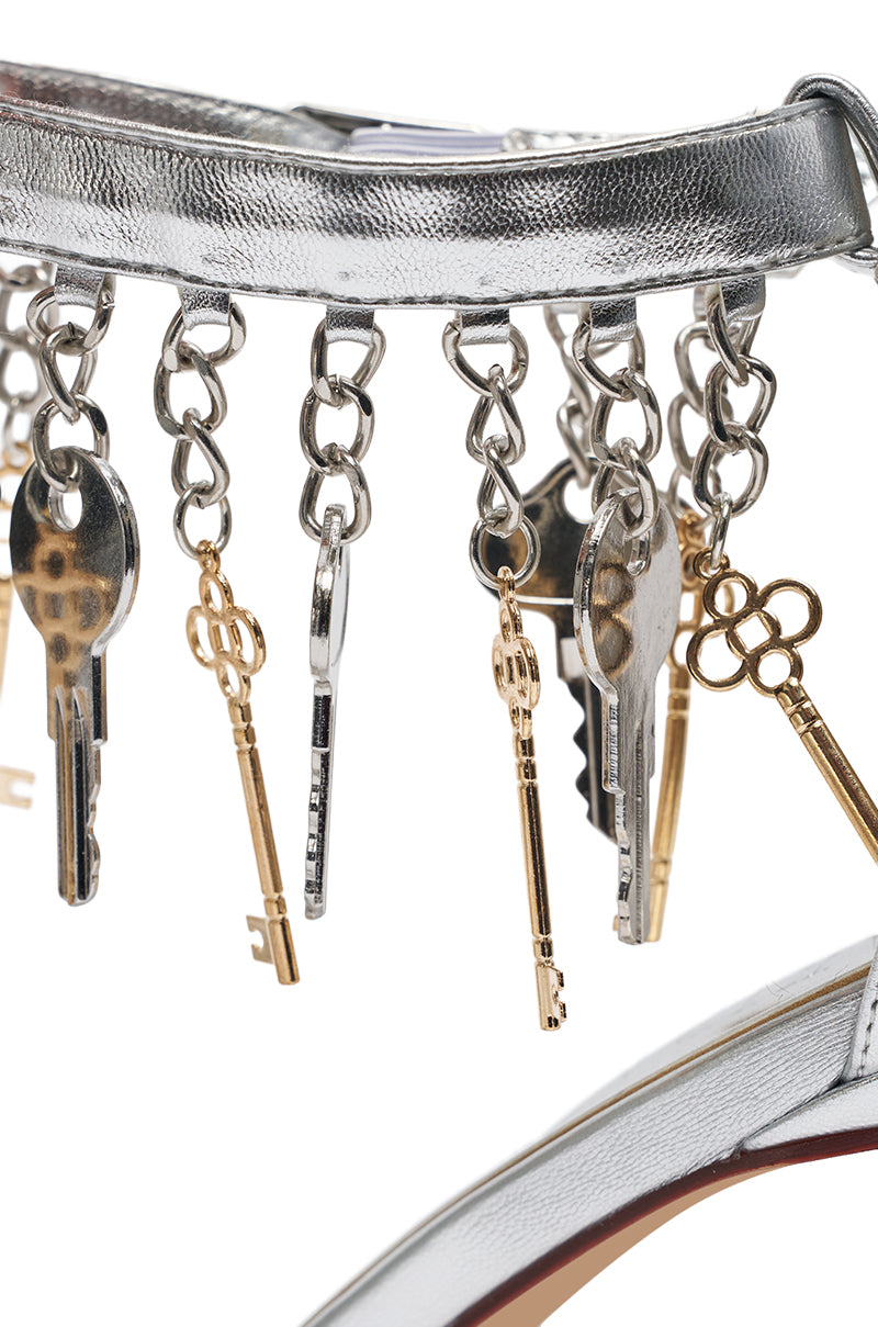 Close-up of the AKIRA GOLD ANNIKI SILVER KEY EMBELLISHED NOVELTY STILETTO SANDAL in metallic faux leather, featuring a strap adorned with silver key charms and short chains in assorted shapes and designs. 8