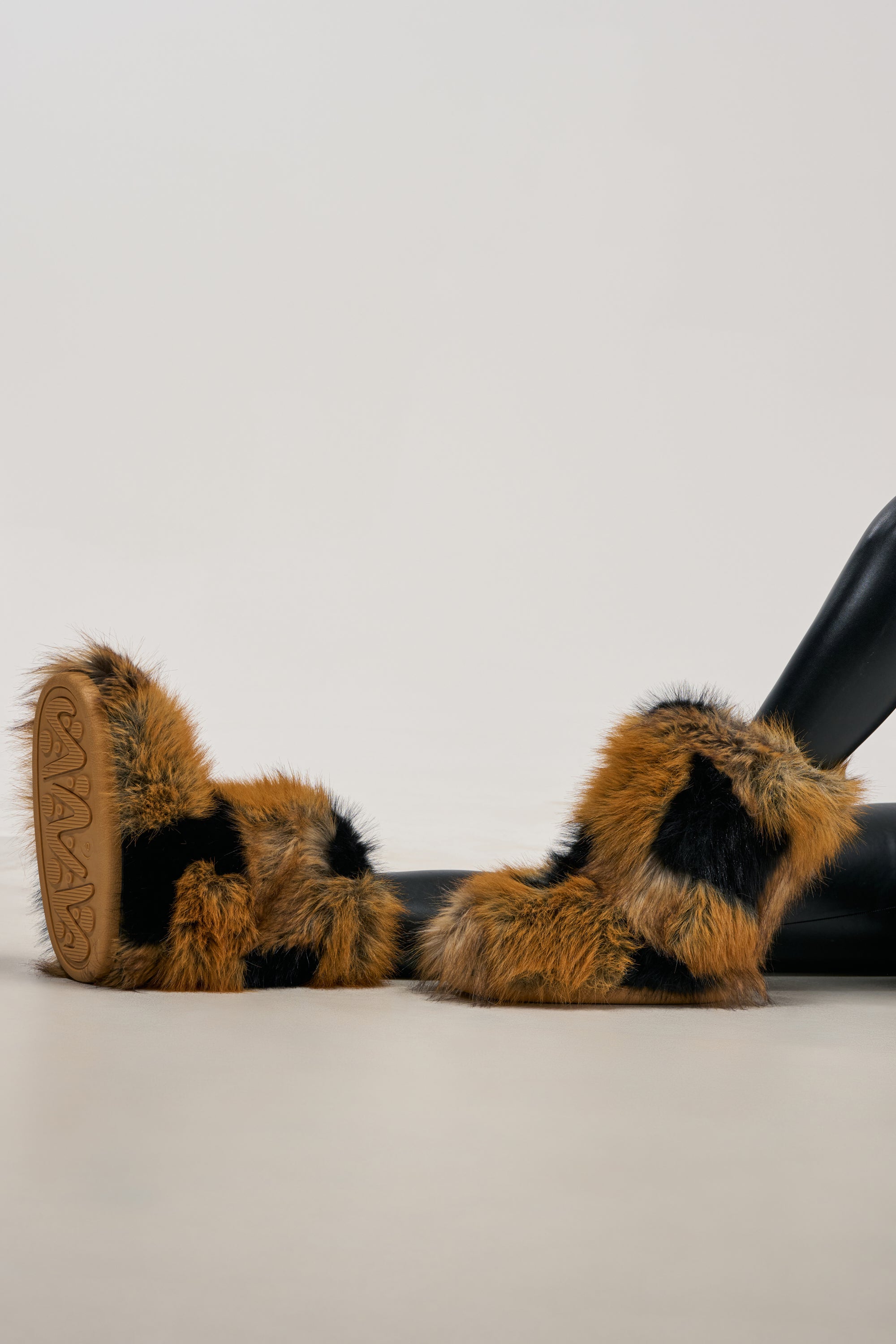 A pair of legs wearing the AZALEA WANG ANSELIE BROWN MULTI PATCHWORK BOOTIE, featuring bold brown and black patchwork faux fur reminiscent of statement streetwear boots, posed against a plain light background.