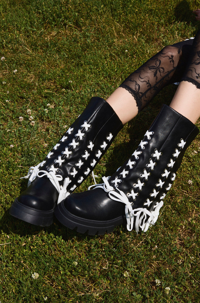 A person wearing the AZALEA WANG ANSELM BLACK LACE TIE BOOTIE with chunky soles and white crisscross laces, paired with black lace tights, sits on grass.