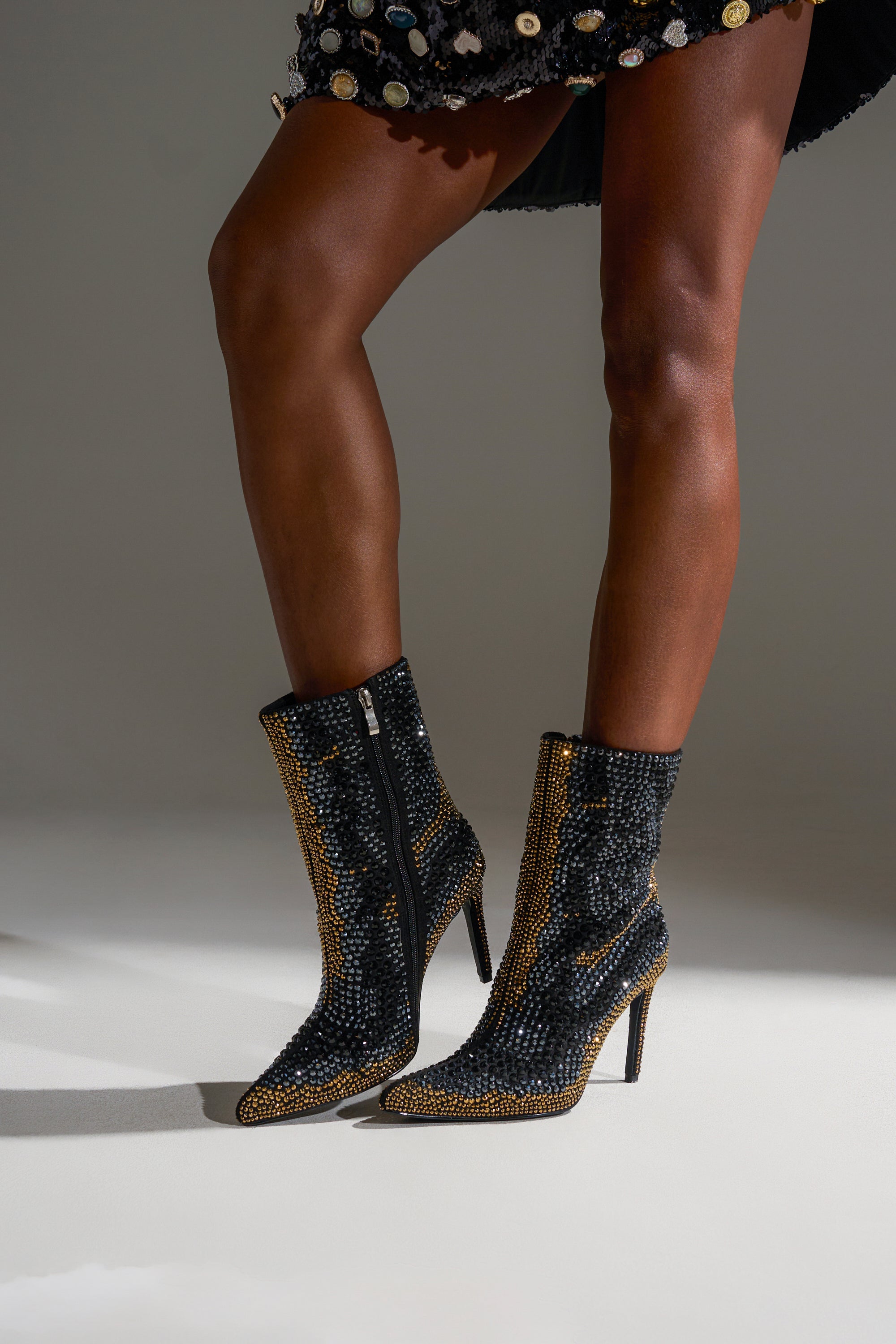 Wearing a black sequin skirt, a person showcases the AZALEA WANG ANSLEY BLACK MULTI RHINESTONE COCKTAIL BOOTIE—sparkly suede ankle boots with gold trim and crystals—while standing on a light surface.