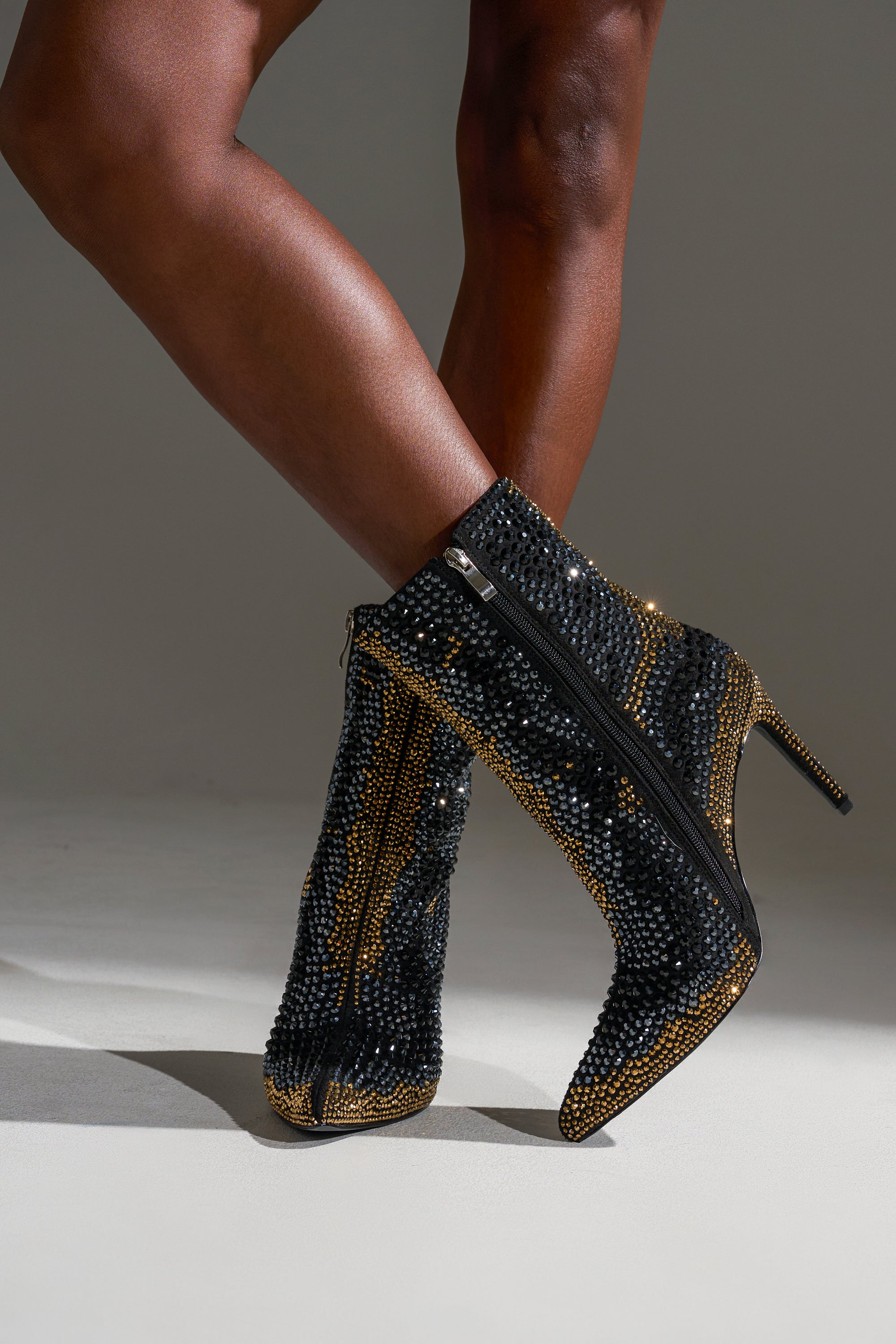 A person with crossed legs wears the AZALEA WANG ANSLEY BLACK MULTI RHINESTONE COCKTAIL BOOTIE—black suede ankle boots with stiletto heels and sparkling gold and rhinestone details—standing on a light-colored surface.