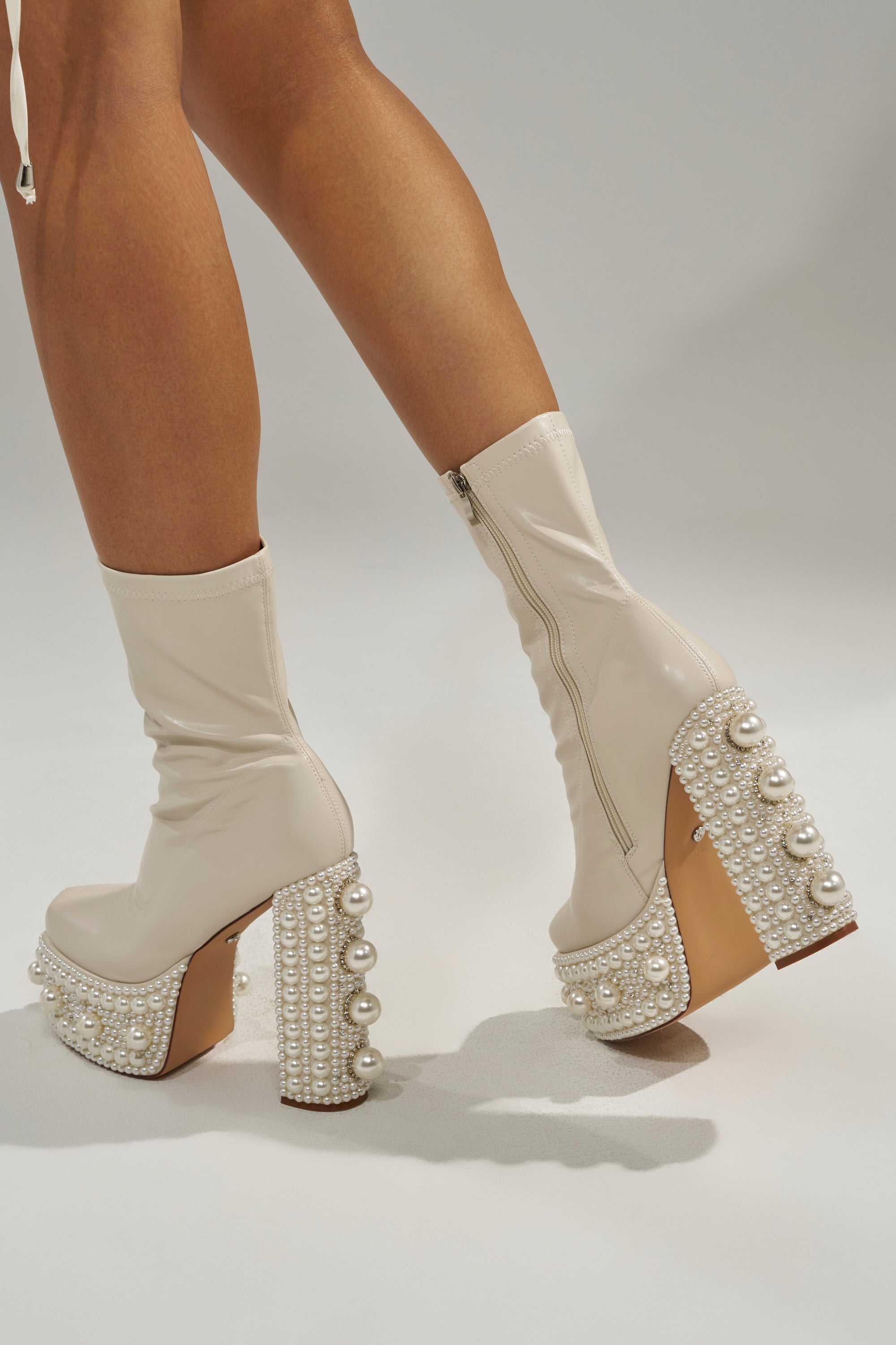 A person wears the AZALEA WANG ANTARIS BEIGE PEARL DETAIL PLATFORM BOOTIE—cream faux leather booties with chunky heels, side zippers, and rows of pearls—standing on a light background with only their legs and feet visible. 0