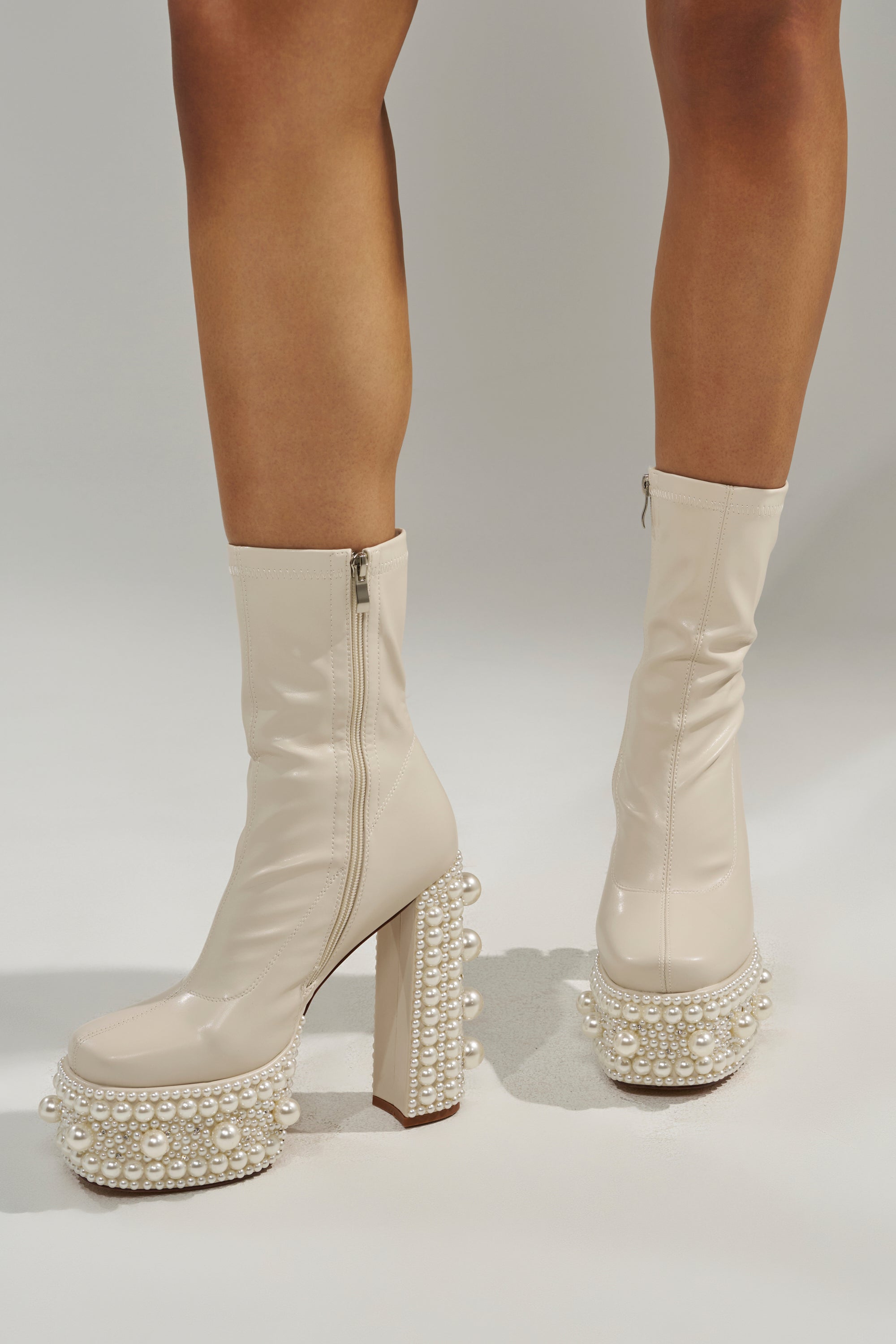 The AZALEA WANG ANTARIS BEIGE PEARL DETAIL PLATFORM BOOTIE features cream faux leather, chunky heels, and platform soles embellished with rows of white pearls, as shown worn on a light-colored surface. 1